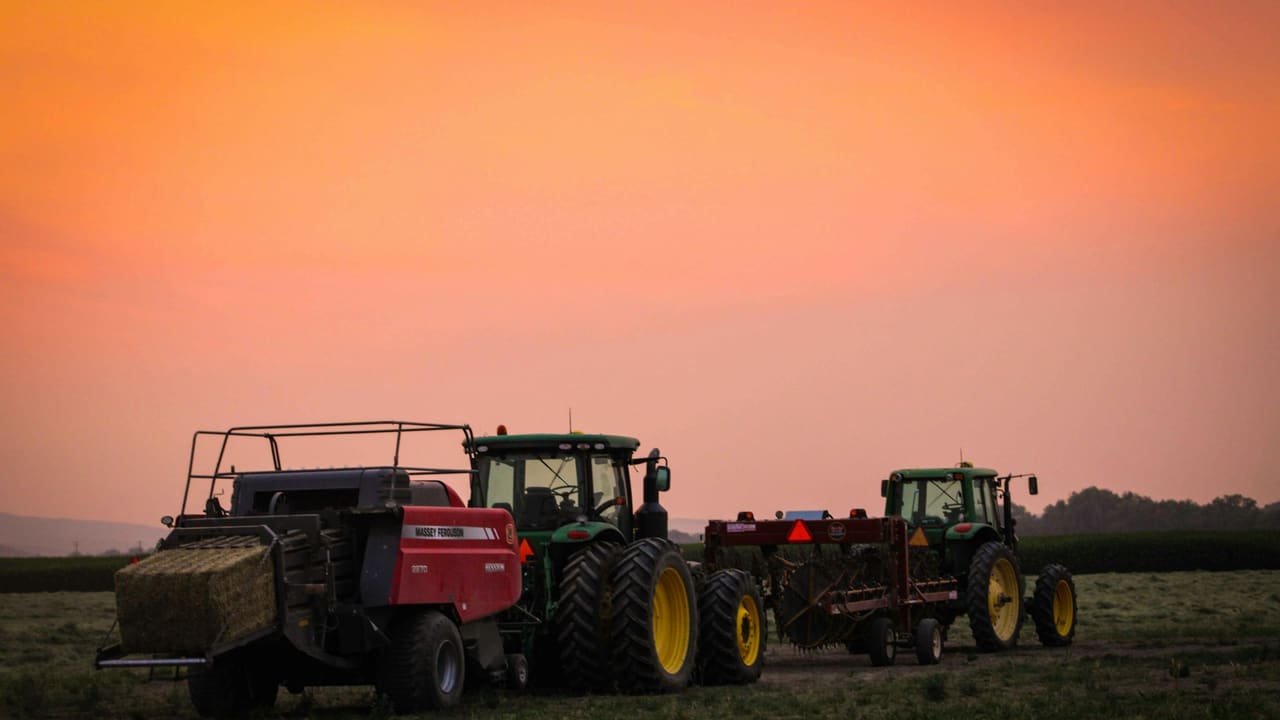 Two tractors and a baler parked at the edge of a mown field at sunset