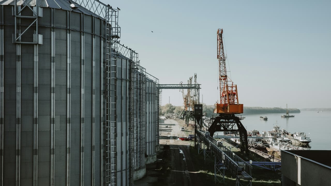 A row of steel grain silos beside a river port, with a loading crane at the quay