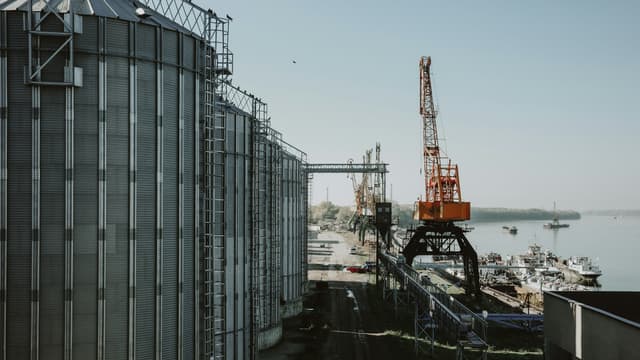 A row of steel grain silos beside a river port, with a loading crane at the quay