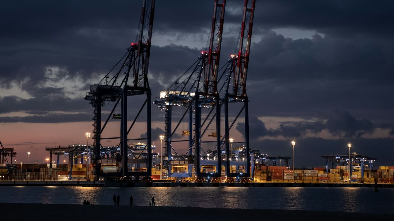 Port cranes standing against a dusk sky, with the lights of a city on the far shore